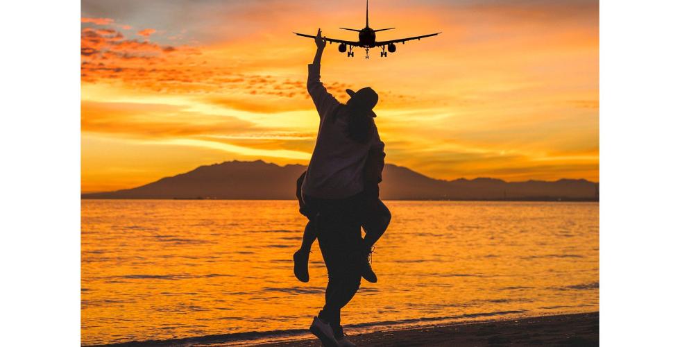 Mann bærer mann på ryggen på en strand i solnedgang med et fly på himmelen.